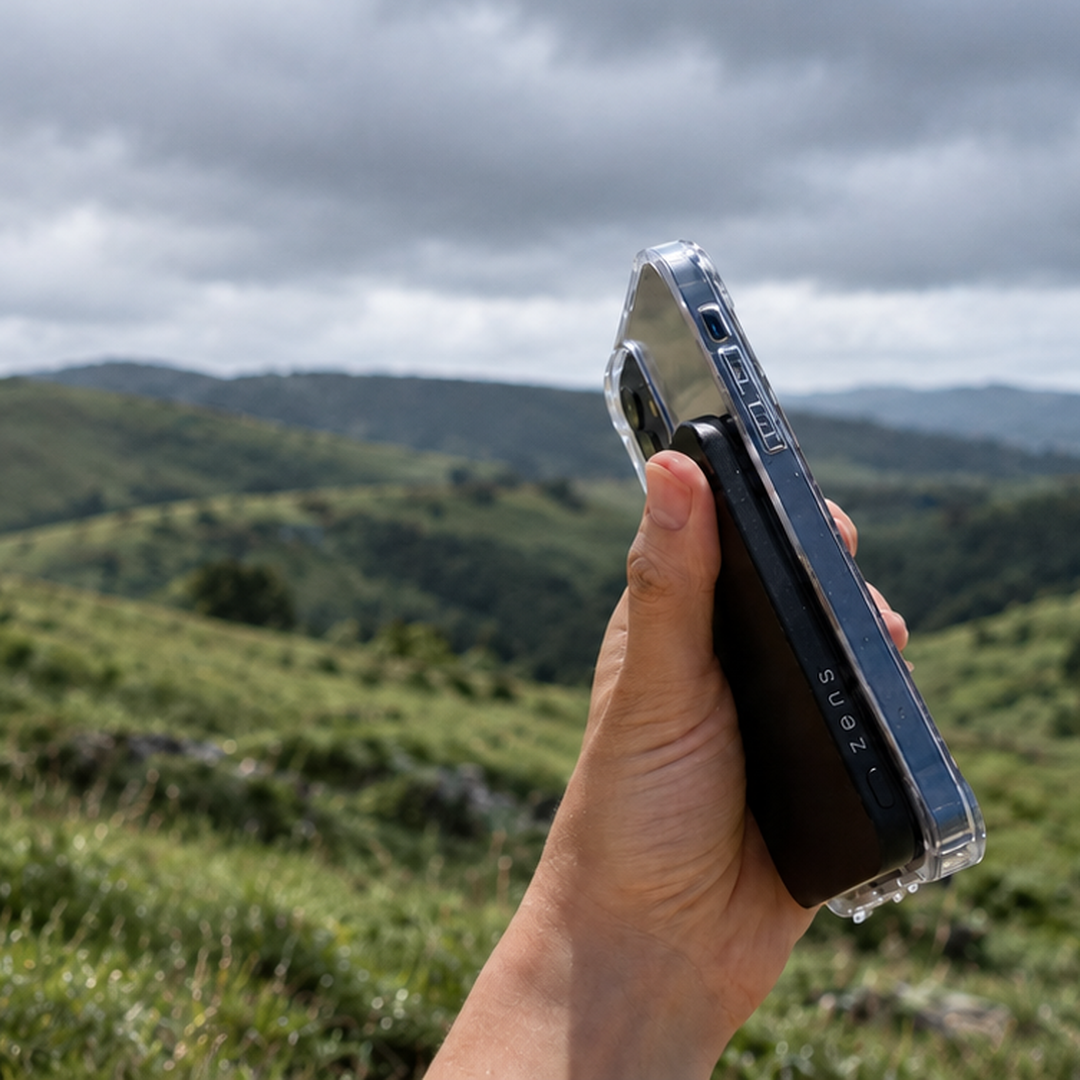A hand holding a black smartphone with Zens powerbank pro 1 in a clear protective case, photographed outdoors against a cloudy sky and green rolling hills.