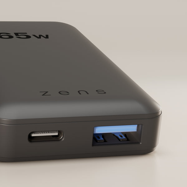 A close-up of a dark gray Zens portable power bank showing its 65W USB-C port and a USB-A port with blue accents, on a neutral light gray background.