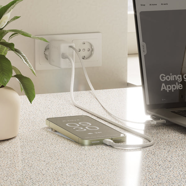 A white USB-C power bank lies on a speckled countertop, connected to a smartphone and also plugged into a USB-C power adapter. A laptop is partially visible on the right, and a potted plant is on the left.