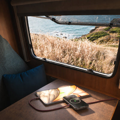 A cozy interior scene of a train or camper with a window showing a coastal landscape outside. On the table in front of the window are a tablet and a smartphone connected to the Zens Charging Cable Pro 2