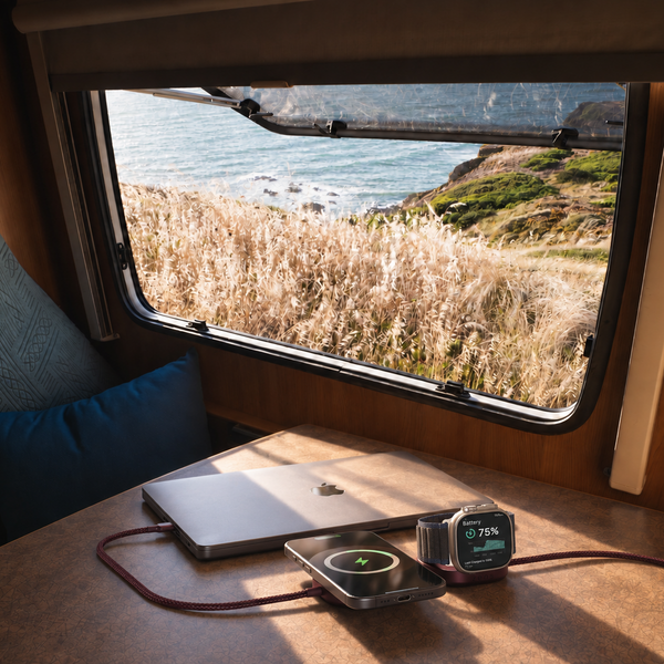 A laptop and a smartwatch are charging on a wooden table inside a camper van, with a scenic view of a coastal cliff and sea visible through the window.