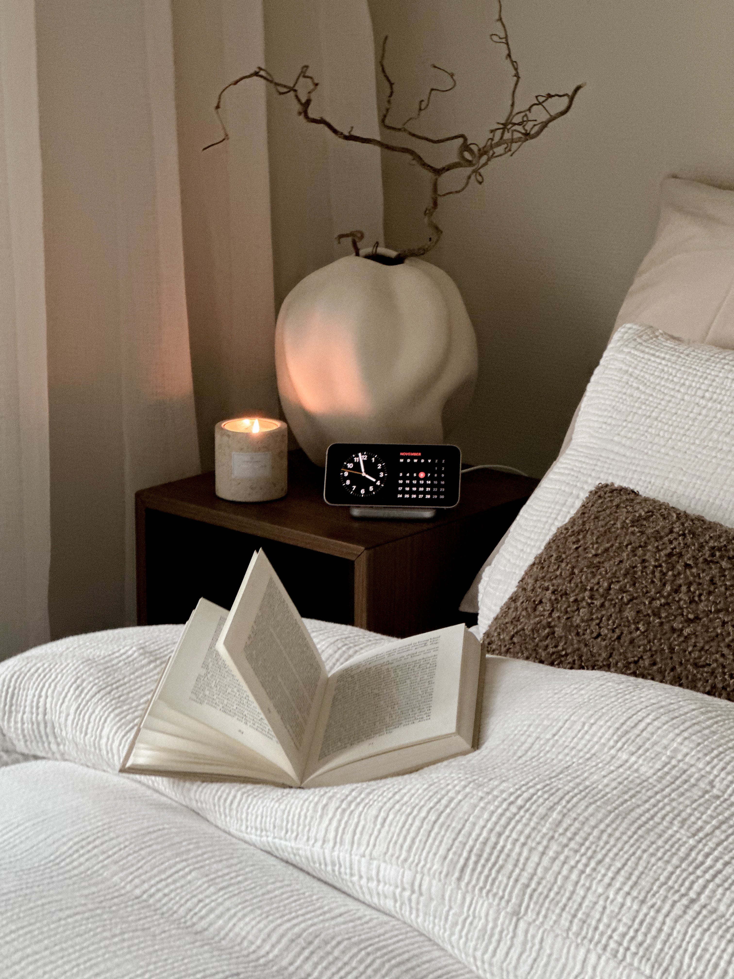 Magnetic Nightstand Charger wirelessly charging a smartphone displaying a clock on a nightstand next to a bed.